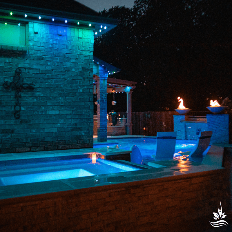 A tranquil night scene unfolds around an inground heated spa, flanked by fire bowls and vibrant colored LED lighting along the edges of the home. In the distance, the patio beckons with its outdoor dining amenities, including a visible green egg cooker. The night glows warmly with the allure of fire and LED features, inviting the family to a cozy and enchanting evening retreat.