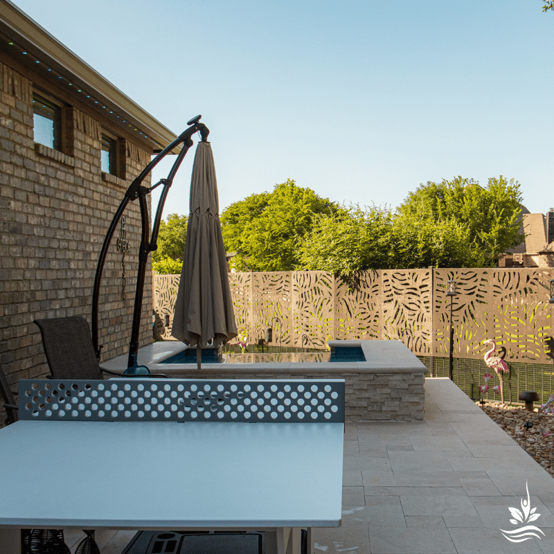A daytime view of an outdoor ping pong table set on richly textured travertine pavers, with a beautiful above-ground heated spa in the background. The spa boasts a 3D depth dry-stacked stone veneer, complementing the lush green trees in the distance, creating a perfect setting for family fun and relaxation.