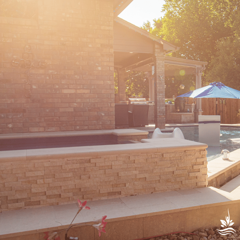 An ethereal daytime view of the backyard, sun-drenched and aglow with lens flares. The family pool and high-end outdoor dining area, equipped with top-tier appliances, bask in the sunlight. A blue pool umbrella unfolds over the tanning ledge, offering a serene shade oasis amidst the vibrant setting.