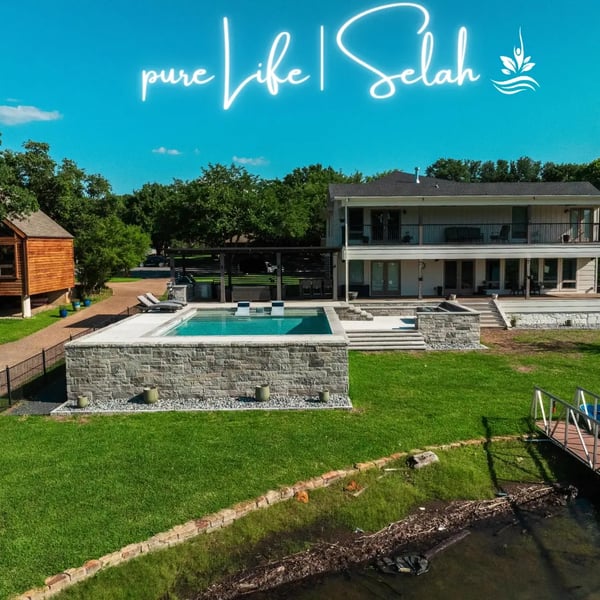 Aerial view of a custom-designed, elevated swimming pool by Selah Pools in Argyle, Texas, featuring multiple levels of steps and walkways. An outdoor dining area under a custom shade structure is visible in the distance, adjacent to the original home. A sunken ‘conversation pit’ area is also included in the design, elevated well above the risk of waterfront encroachment.